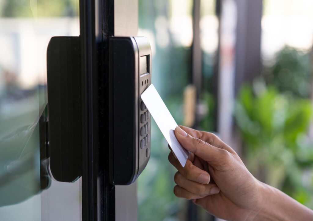 A hand holds a white key card near a black card reader on a glass door, suggesting entry access. Background shows blurred greenery.