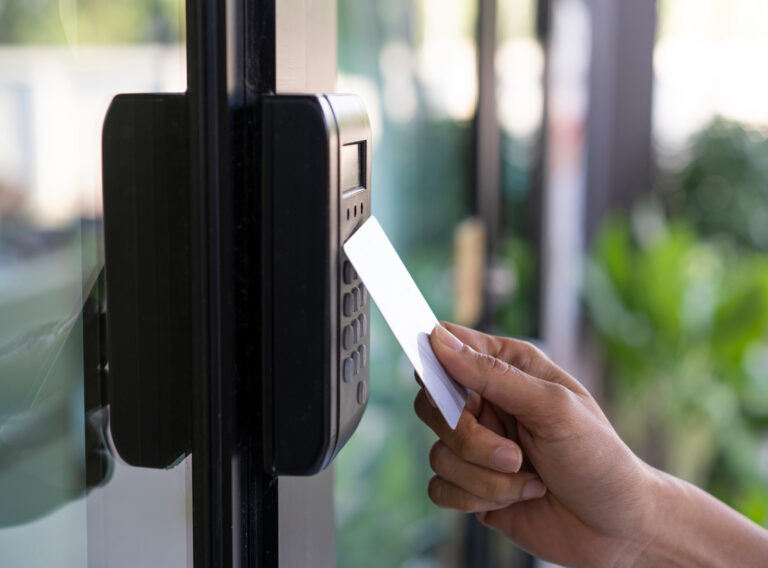 A hand holds a white key card near a black card reader on a glass door, suggesting entry access. Background shows blurred greenery.
