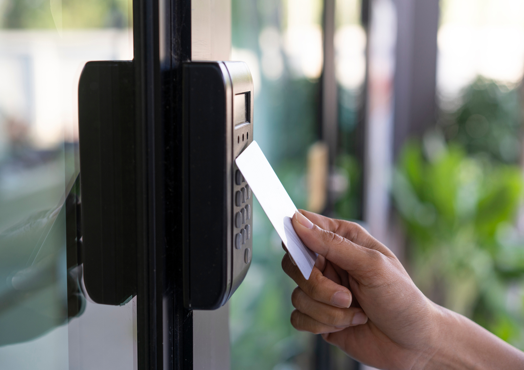 Enhance Your Business Security with a Professional Access Control Systems 8 A hand holds a white key card near a black card reader on a glass door, suggesting entry access. Background shows blurred greenery.